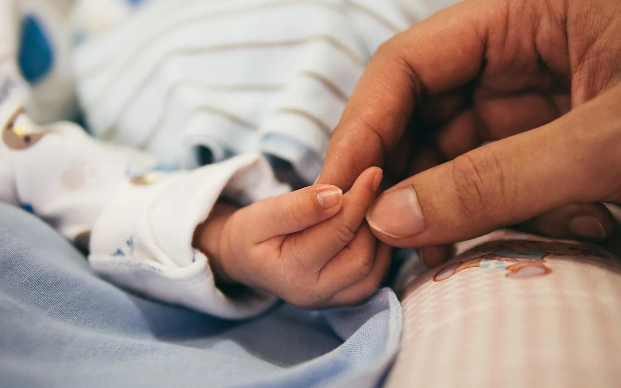 Main d'un adulte qui effleure la minuscule main d'un nouveau-né, vêtu d'un body rayé, sur des draps bleu clair